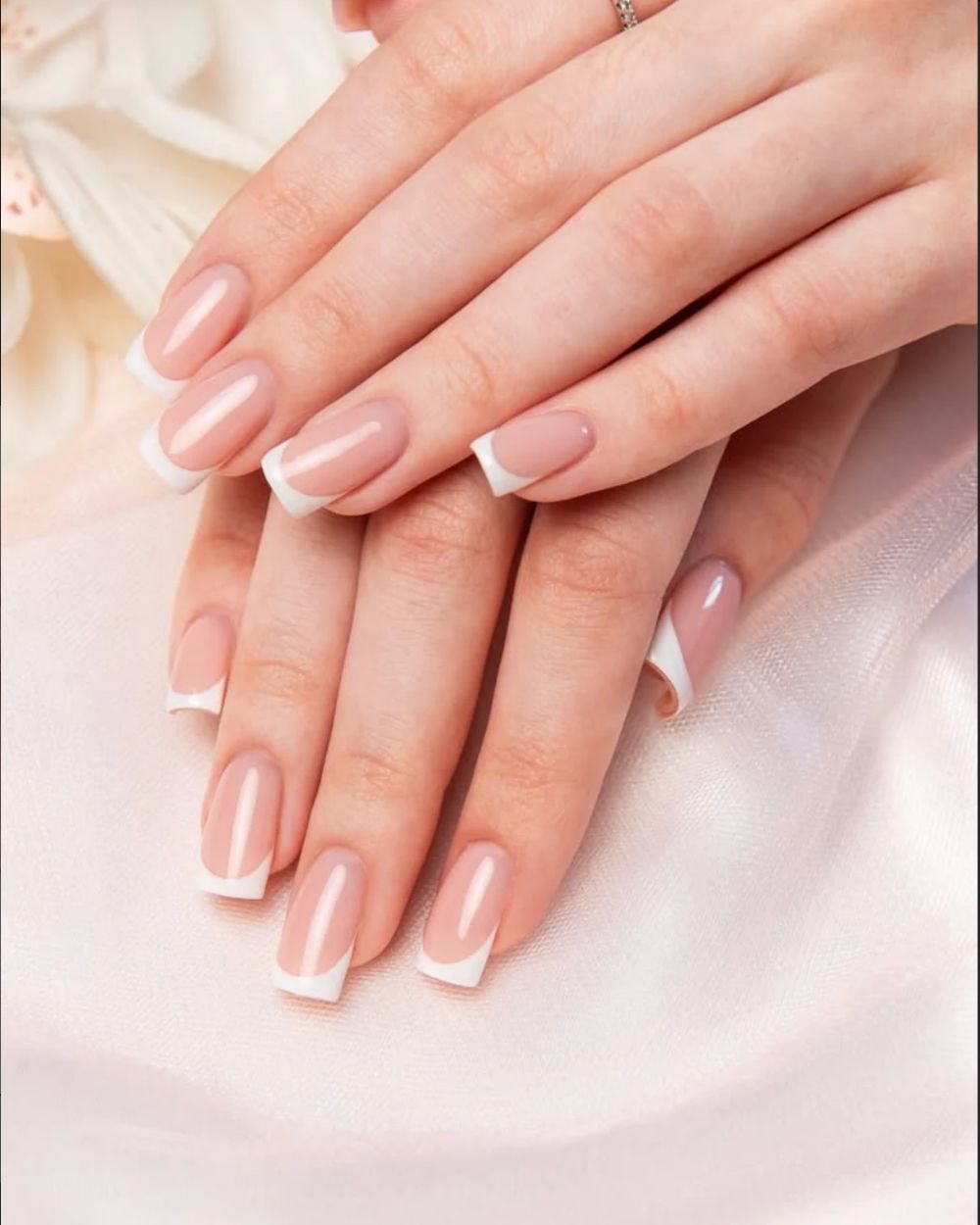 Two hands resting on cream silk, long square French tips with a nude bed and a white edge.