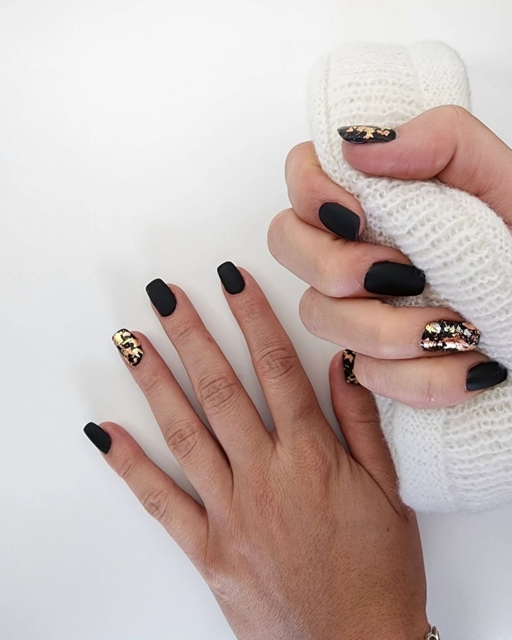 A hand against a white knit sweater, matte black nails with gold foil worked across two of them.