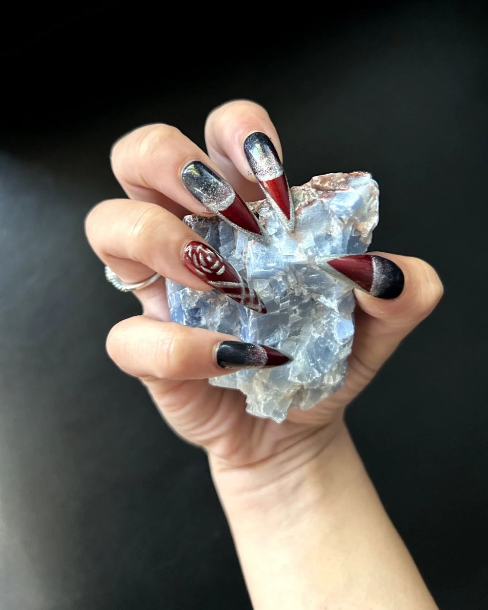 A hand holding a rough blue calcite stone, long stiletto nails in oxblood red, black and chrome.